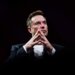 Elon Musk seated with hands clasped, wearing a suit and looking thoughtfully ahead against a dark background.