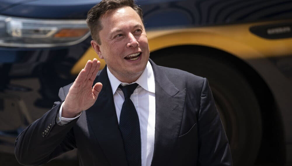 Elon Musk waving while wearing a suit, with a vehicle in the background.