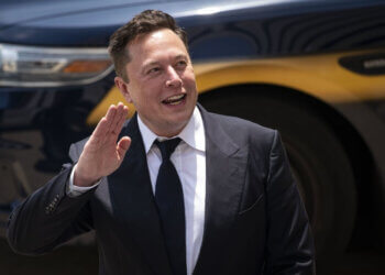 Elon Musk waving while wearing a suit, with a vehicle in the background.