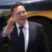 Elon Musk waving while wearing a suit, with a vehicle in the background.