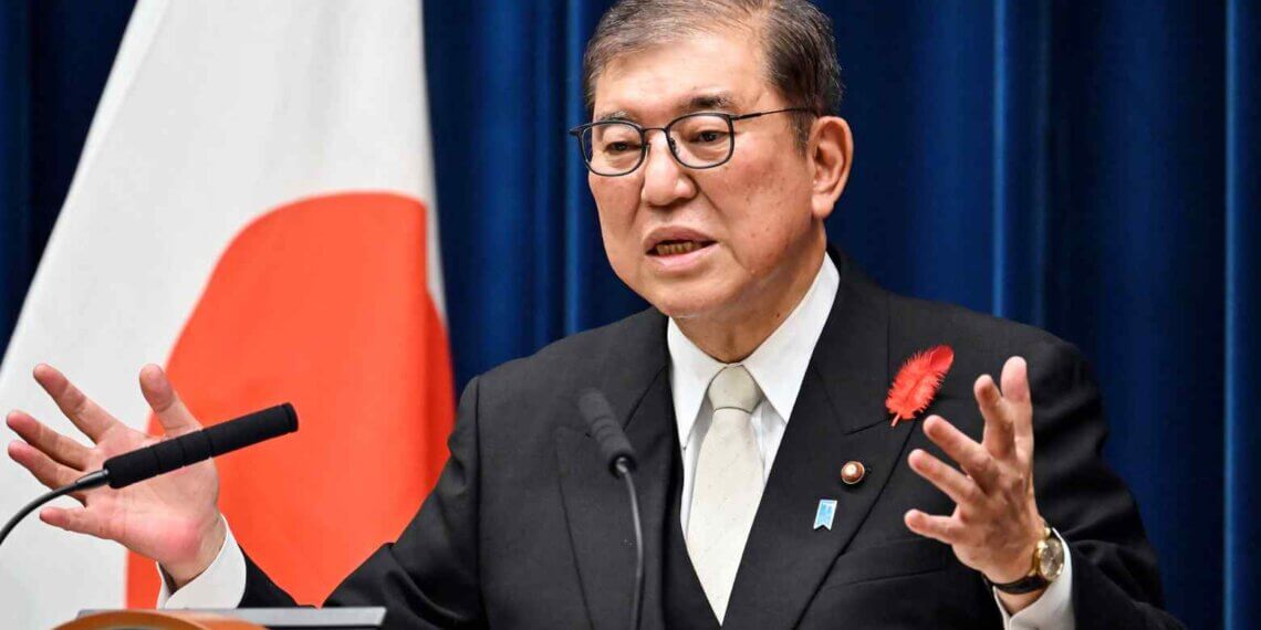 A government official in a suit speaking at a podium with a Japanese flag in the background.