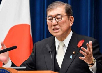 A government official in a suit speaking at a podium with a Japanese flag in the background.
