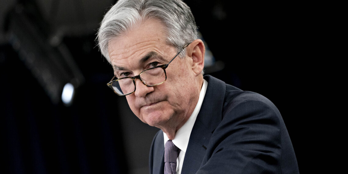 Jerome Powell adjusting his glasses during a press conference, wearing a dark suit and tie.