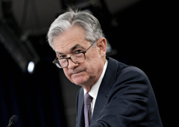 Jerome Powell adjusting his glasses during a press conference, wearing a dark suit and tie.