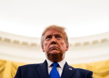 Donald Trump wearing a dark suit and blue tie, looking serious in a formal setting.