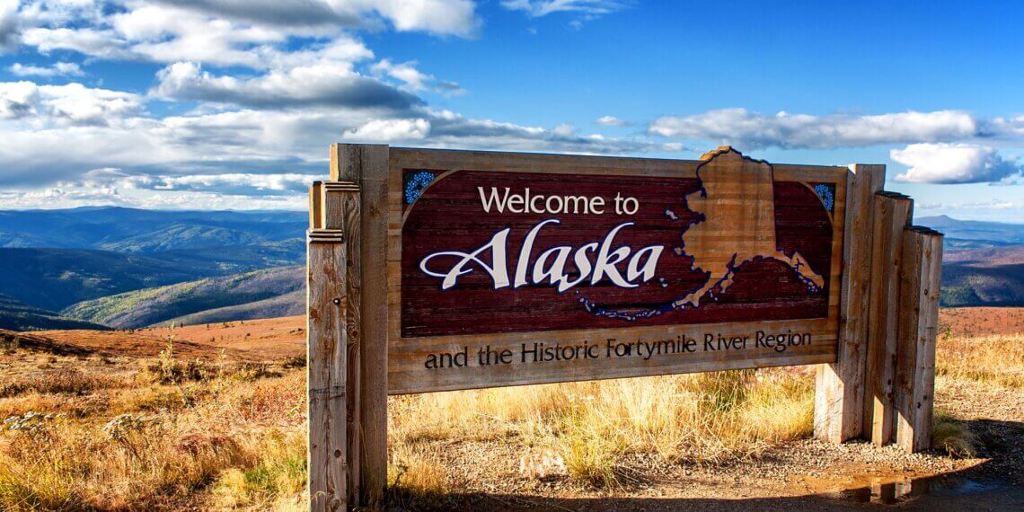 Wooden sign that reads 'Welcome to Alaska' with a map of Alaska and scenic background.