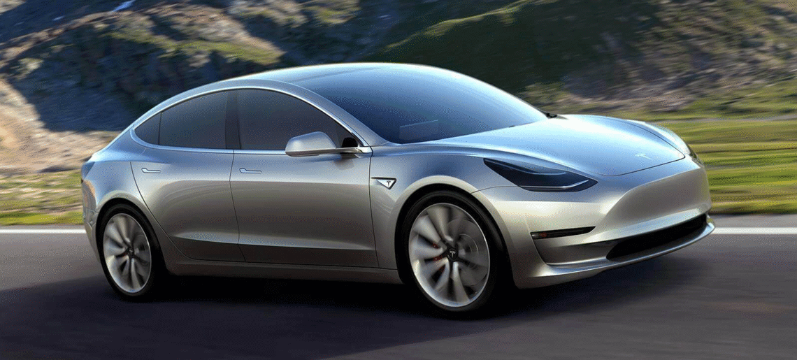 Silver Tesla Model 3 electric car driving on a road with a mountainous background.