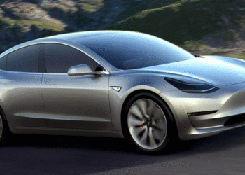 Silver Tesla Model 3 electric car driving on a road with a mountainous background.