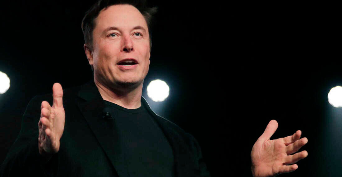 A man in a black suit gesturing while speaking on stage with lights in the background.