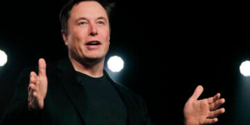 A man in a black suit gesturing while speaking on stage with lights in the background.