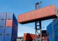 A forklift lifting a red shipping container in a cargo yard surrounded by stacked blue containers.