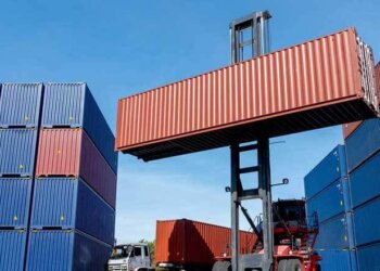A forklift lifting a red shipping container in a cargo yard surrounded by stacked blue containers.