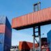 A forklift lifting a red shipping container in a cargo yard surrounded by stacked blue containers.